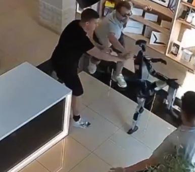 Кадър от охранителна камера, показващ хуманоиден робот в бойна поза срещу клиент и служители в магазин за електроника.