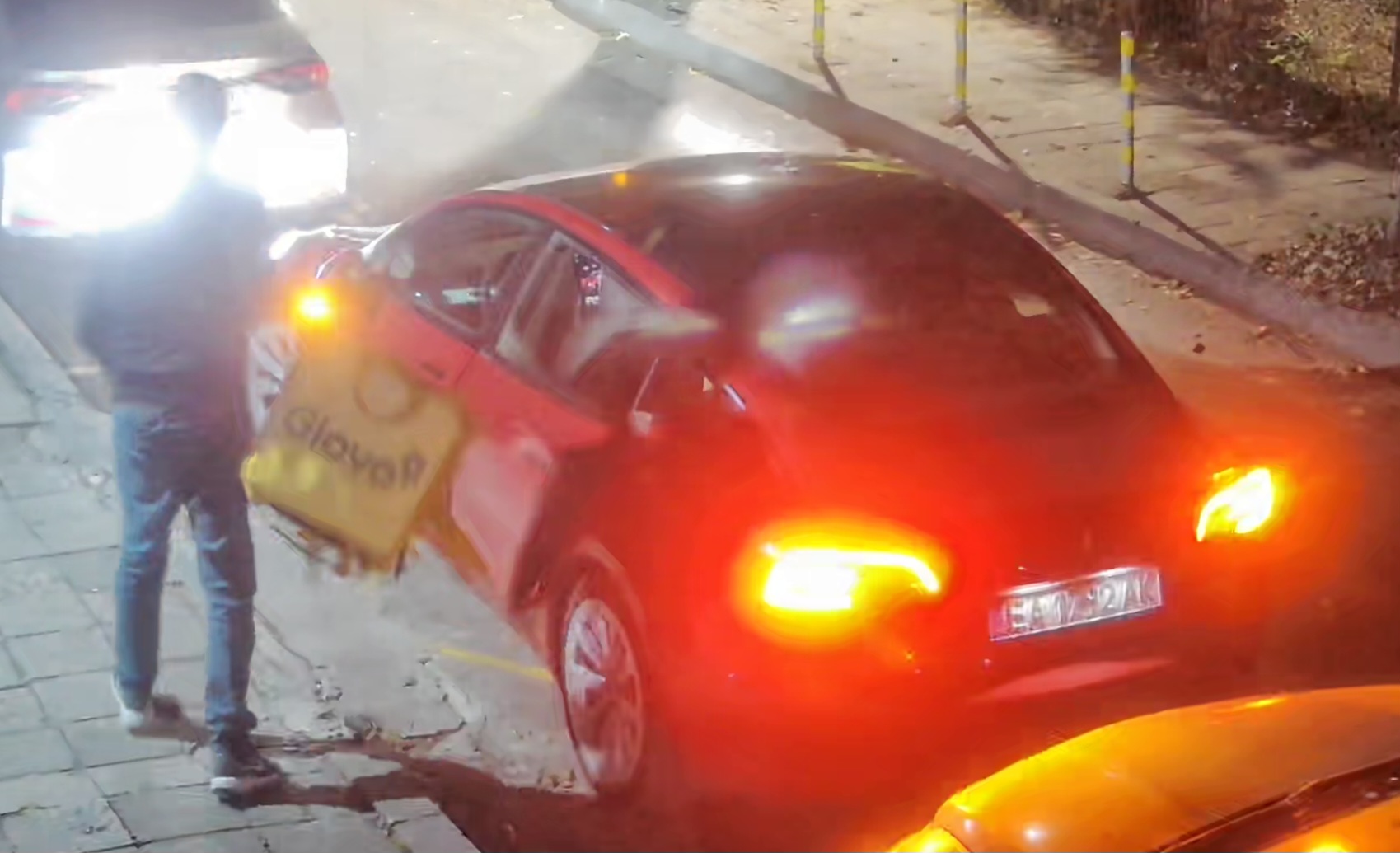 Нощна снимка на червена Tesla Model 3 на пътя, от която доставчик изважда жълта термокутия за храна с логото на популярна фирма.